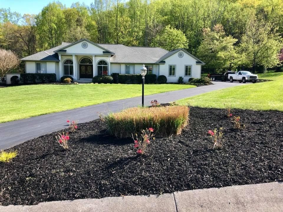 Single-story white house with a black driveway, landscaped flower bed, and trees in the background