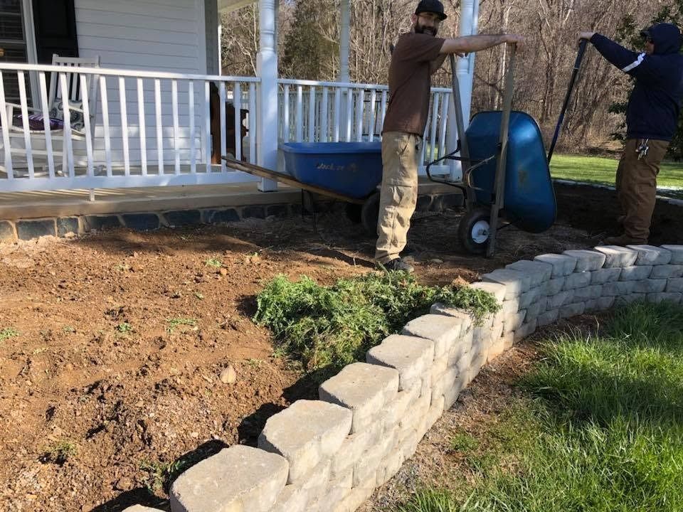 Two workers install a white railing beside a house, with a stone retaining wall and dirt yard.