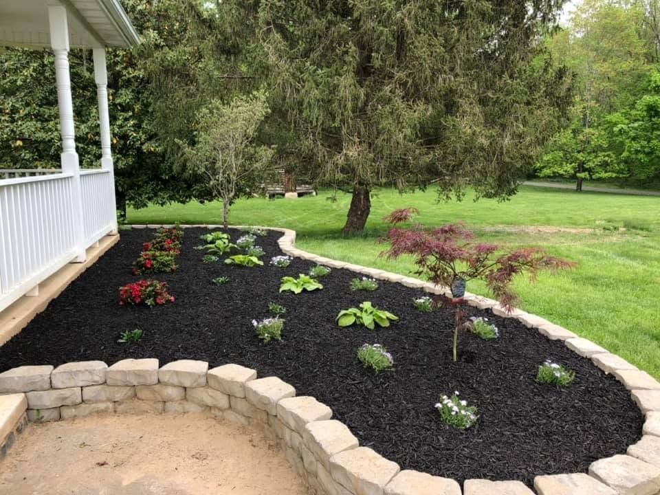 Curved flower bed with black mulch and small plants beside a porch, bordered by tan pavers on a green lawn
