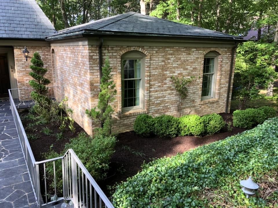 Stone house corner with arched windows, white railing, and landscaped garden in a wooded setting