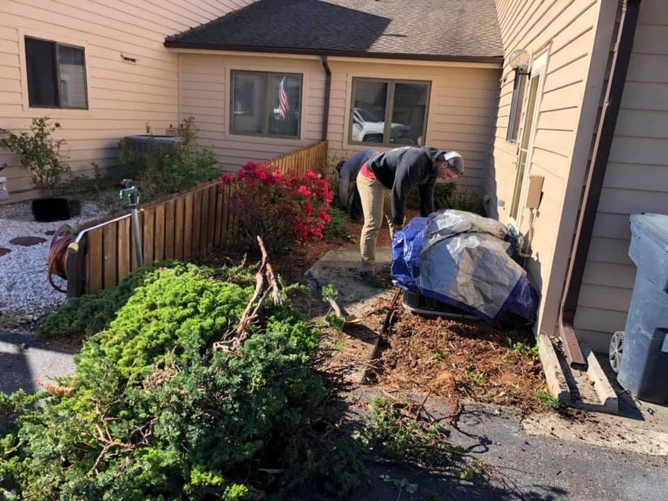 Two people cleaning up yard debris beside a house, with branches, a tarp, and a pile of mulch.