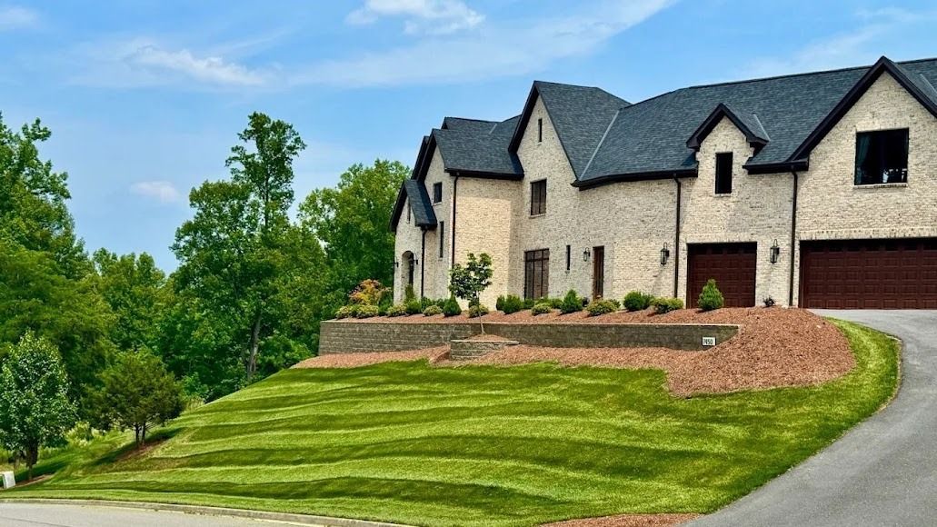Large white house with dark roof on a grassy hillside beside a curved driveway and trees