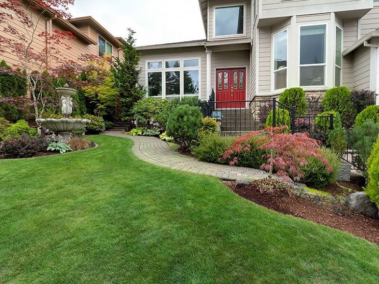 Front yard garden with lush green lawn, curved walkway, shrubs, and a house with a red front door