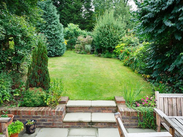Garden lawn with stone steps, brick edging, and lush green shrubs around a patio seating area