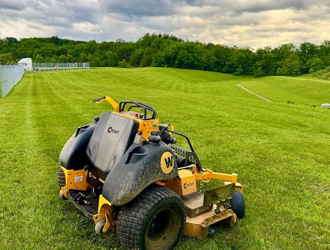 Yellow lawn mower on a grassy field near a tree line under cloudy skies