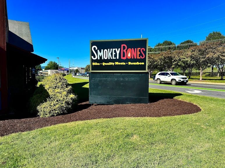 Smokey Bones restaurant sign by a road with lawn, shrubs, and a white SUV in the background