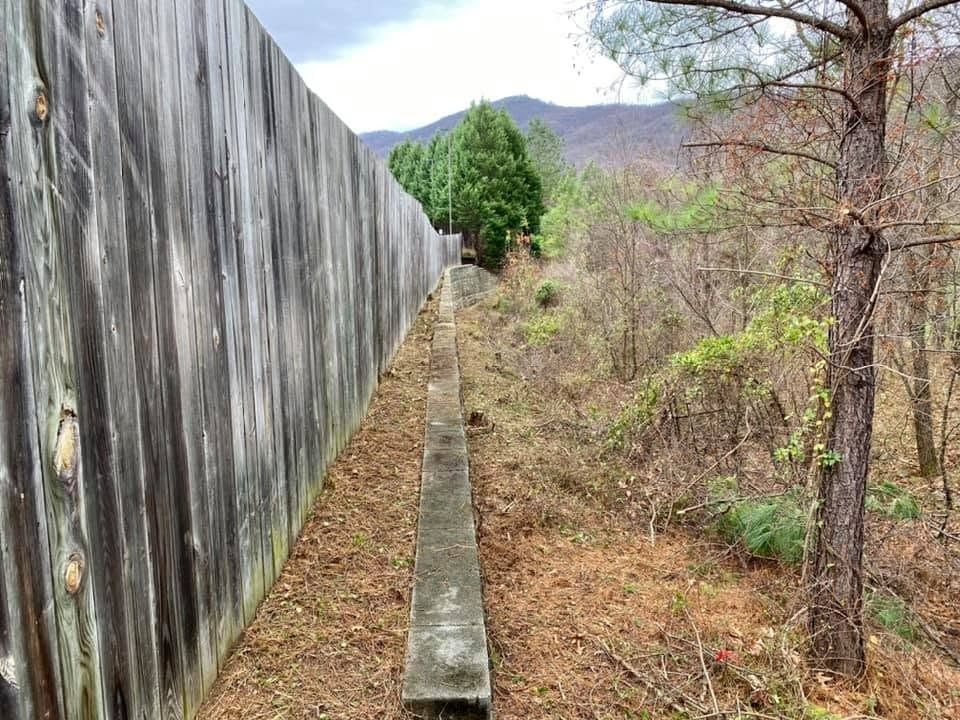 Narrow concrete ledge beside a tall wooden fence in a dry, wooded area with mountains in the background