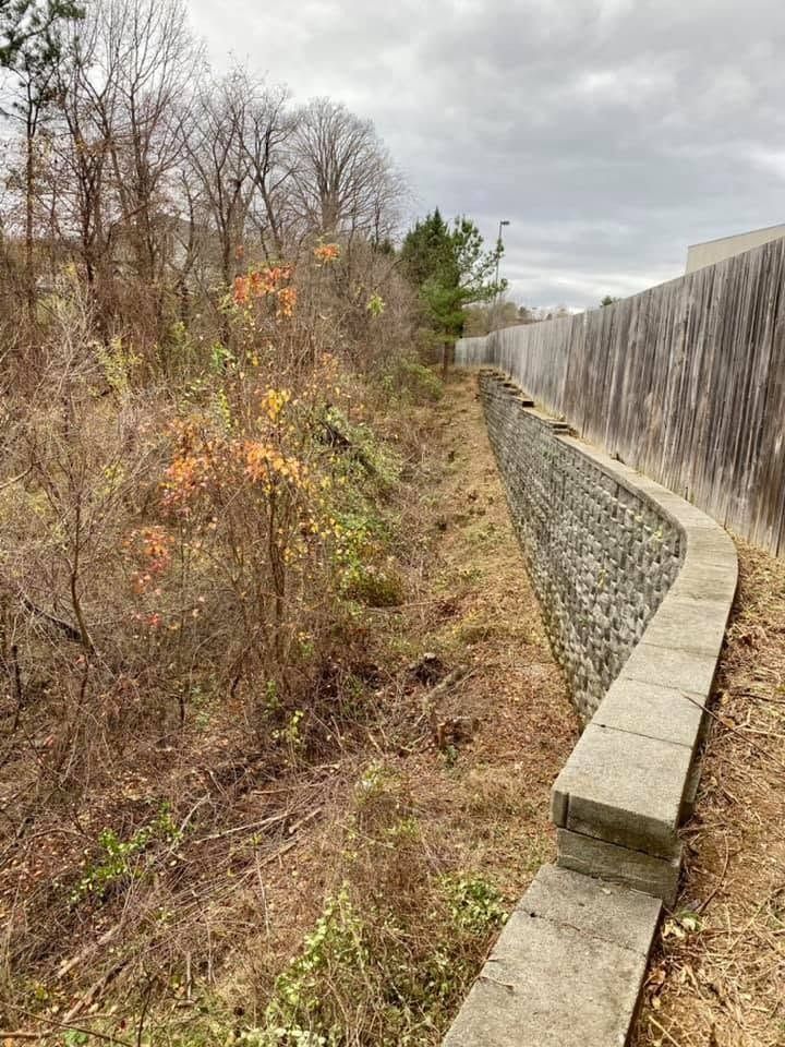 Drainage channel beside a retaining wall along a wooded hillside under a cloudy sky