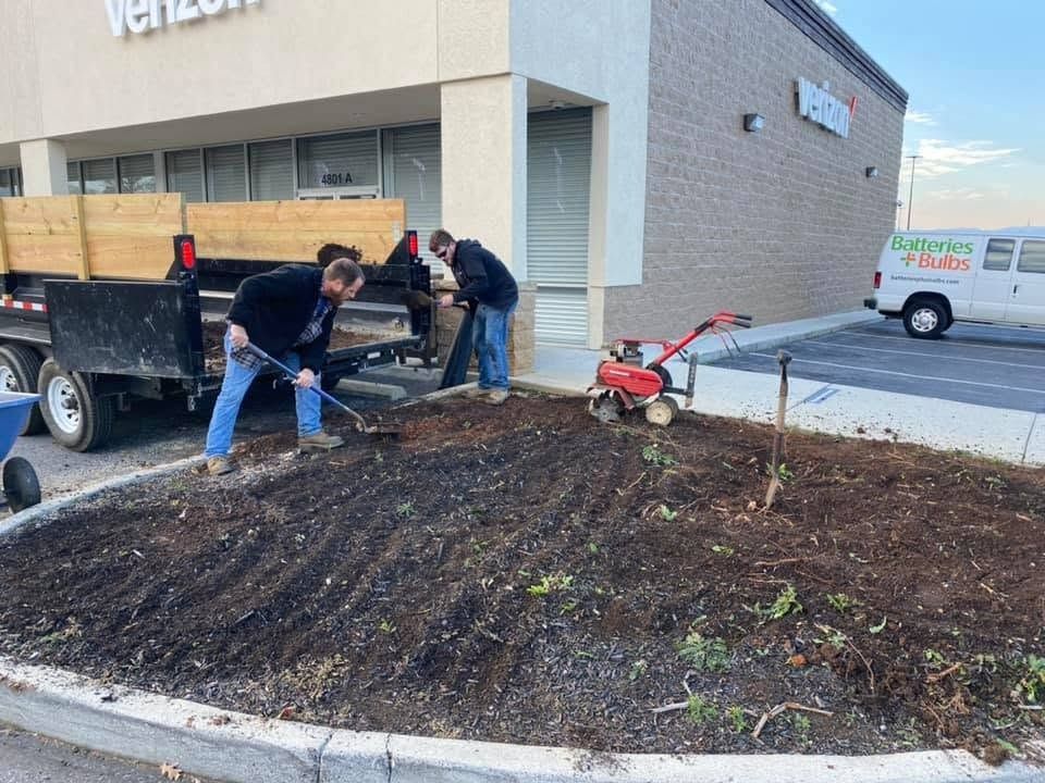 Workers spreading mulch beside a building, with a wheelbarrow and red tiller in the foreground.