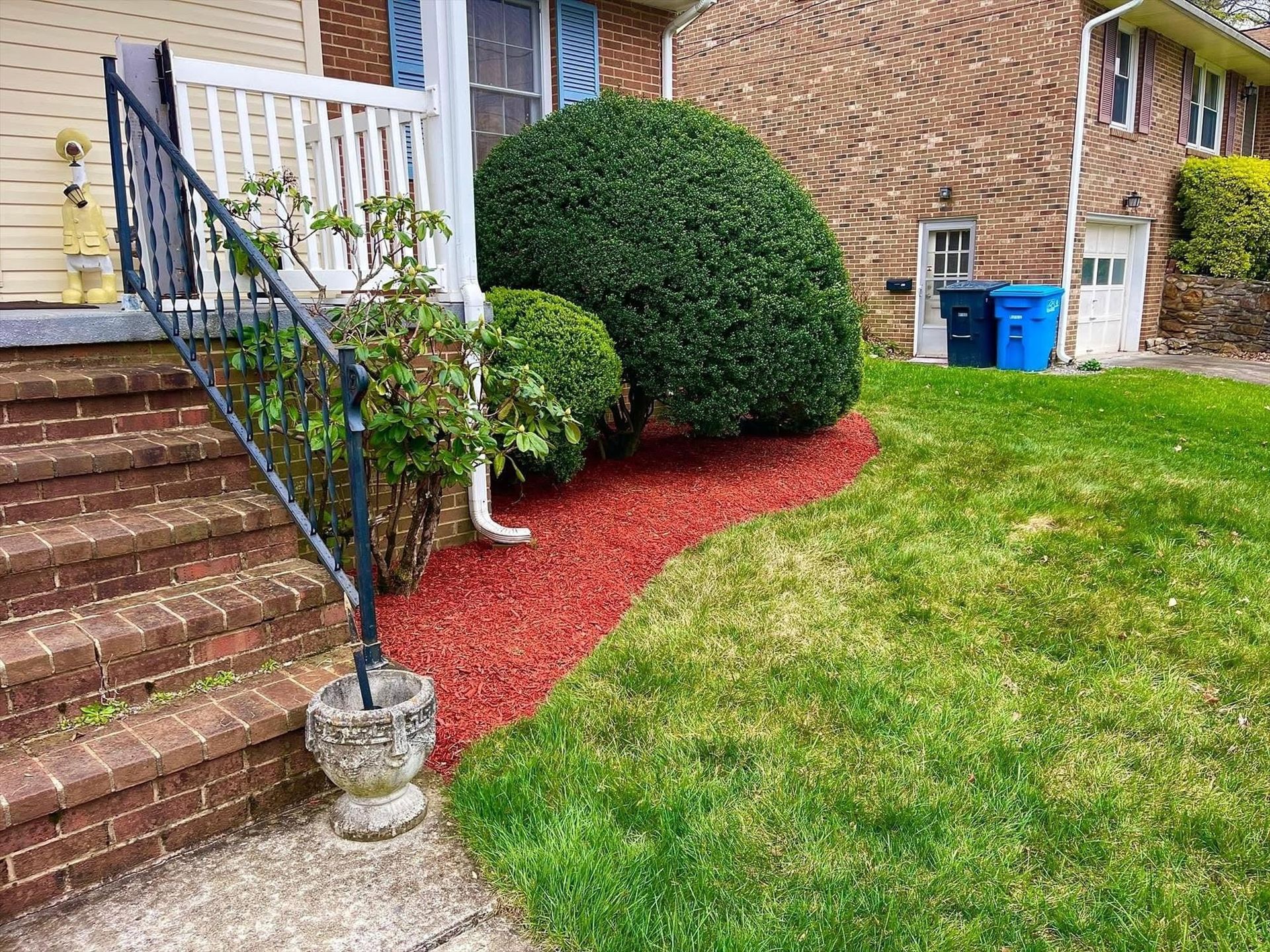 Front yard with brick steps, trimmed bushes, red mulch, and green lawn beside a house.