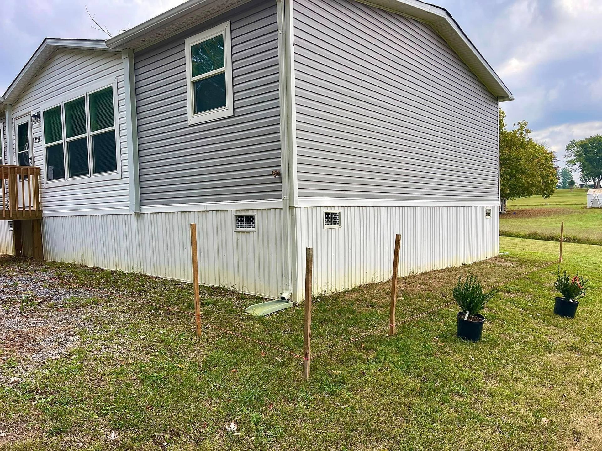 White mobile home with gray siding and white skirting on a grassy lot with young stakes and small potted plants.