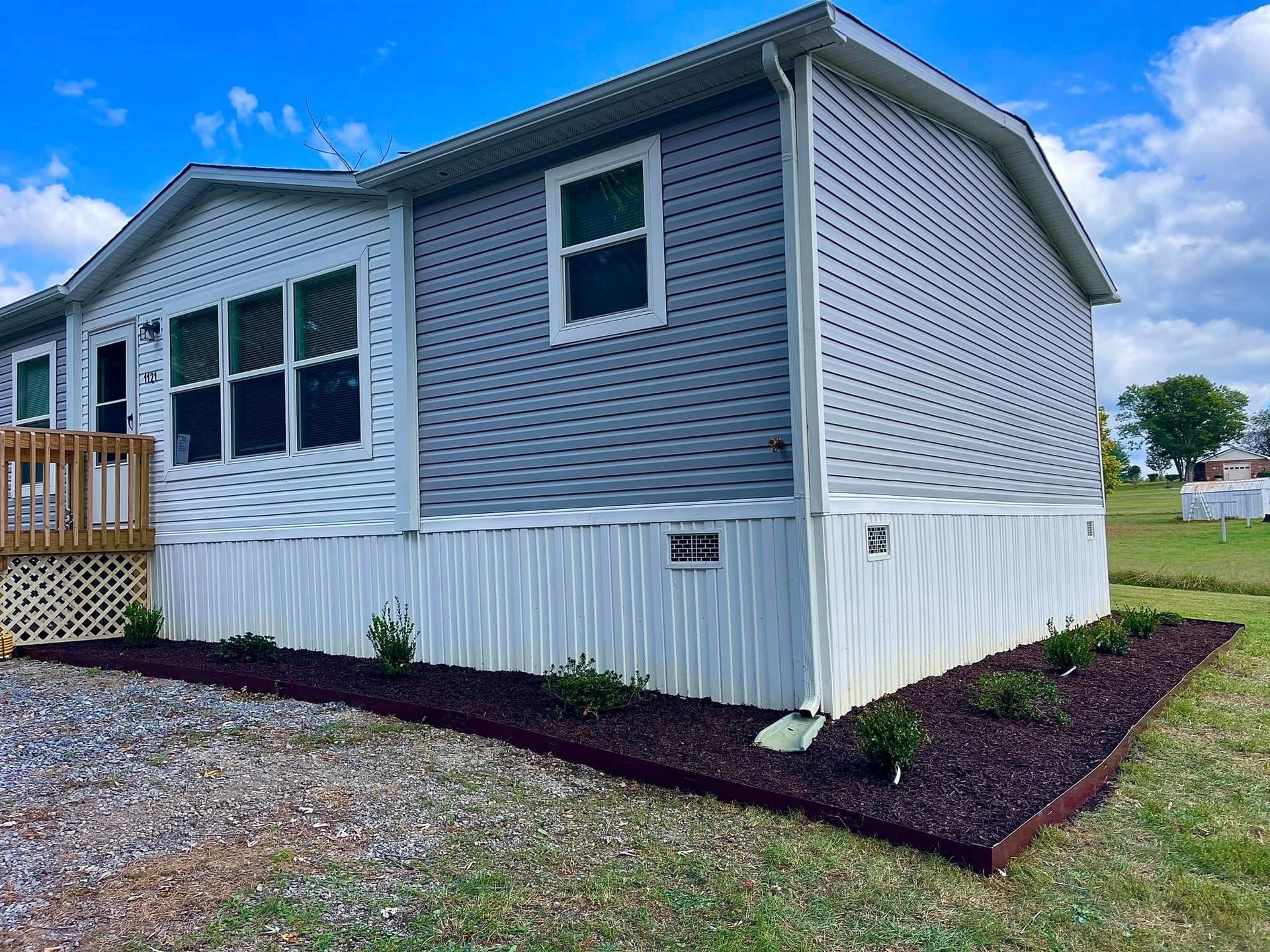 Gray manufactured home with white trim and skirting, gravel yard, and mulched flower bed under a blue sky