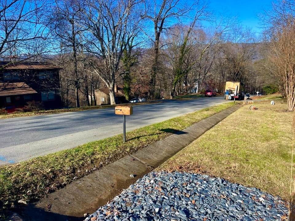 Quiet rural road beside a grassy ditch, leafless trees, and a small sign under a blue sky.