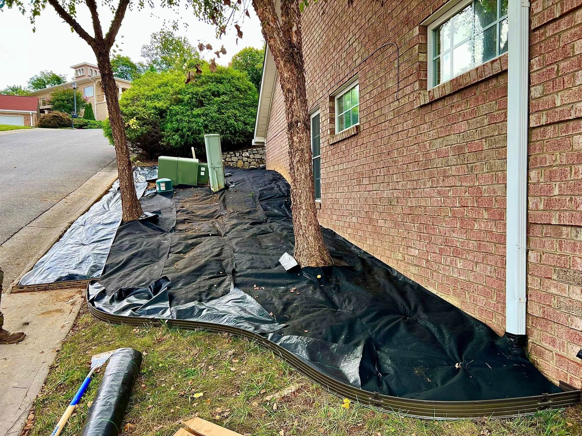 Black tarp laid along a brick house foundation beside a sidewalk and tree, with green equipment nearby