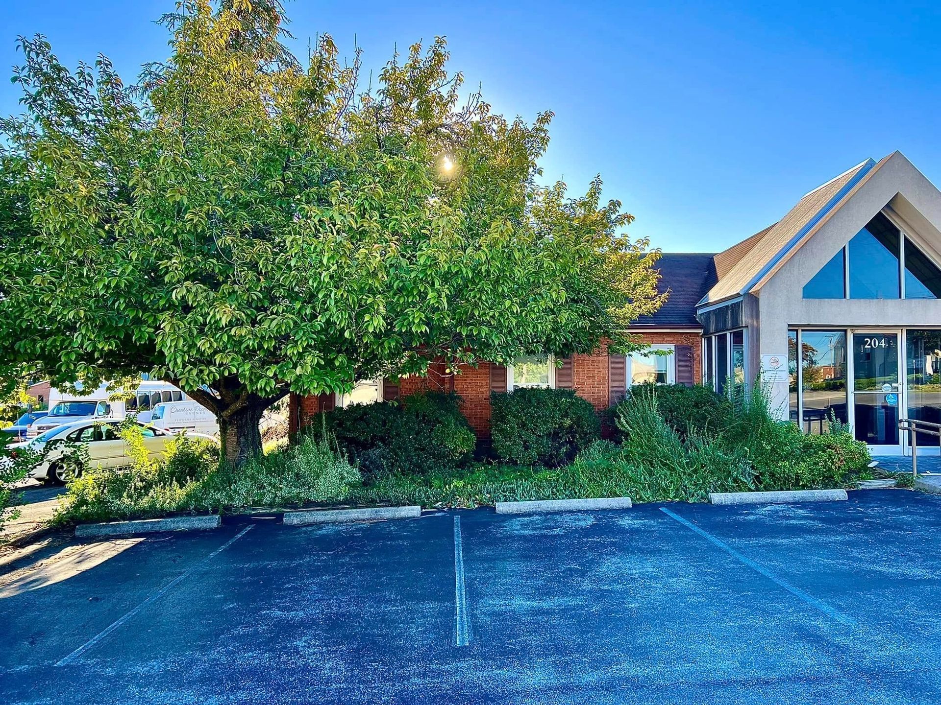 Small brick building with a glass entrance, surrounded by trees and shrubs beside an empty parking lot.
