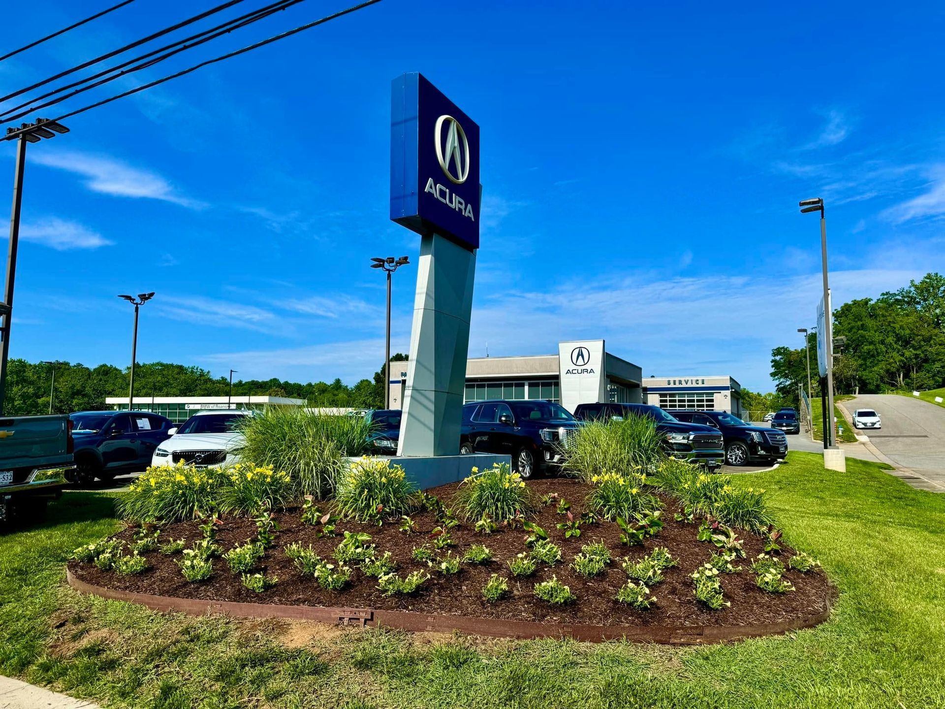 Acura dealership sign in a landscaped lot with parked cars and blue sky