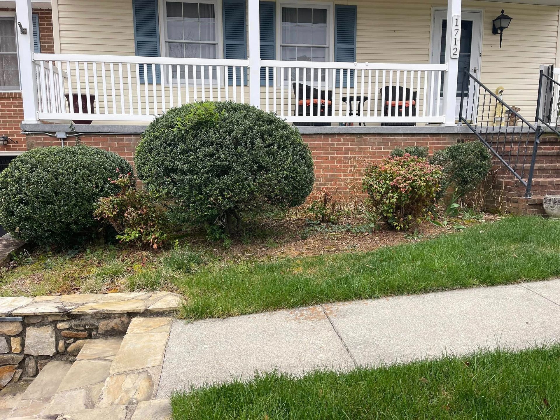 Front porch of a house with white railing, brick foundation, trimmed shrubs, and a sidewalk in front.
