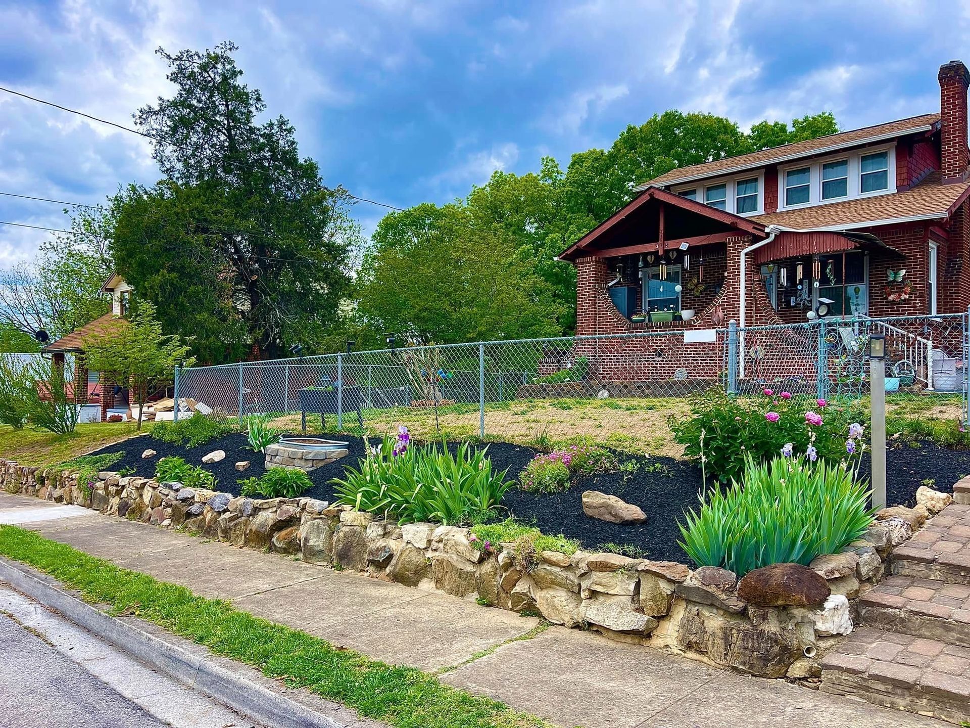 Brick house with a front garden, stone retaining wall, and leafy trees beside a quiet street