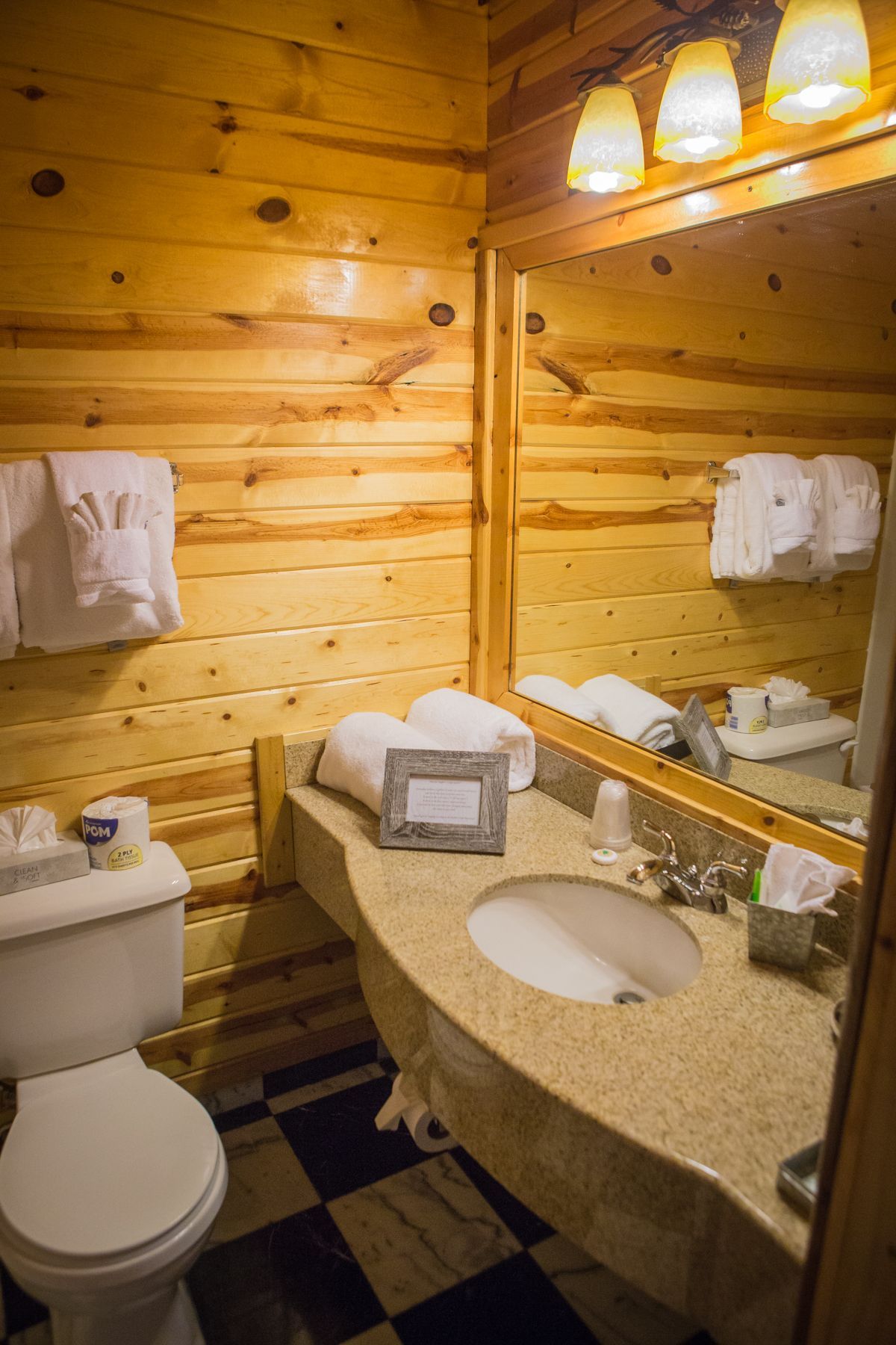 Bathroom with wood paneling, granite countertop, mirror, toilet, and towels. Black and white tiled floor.