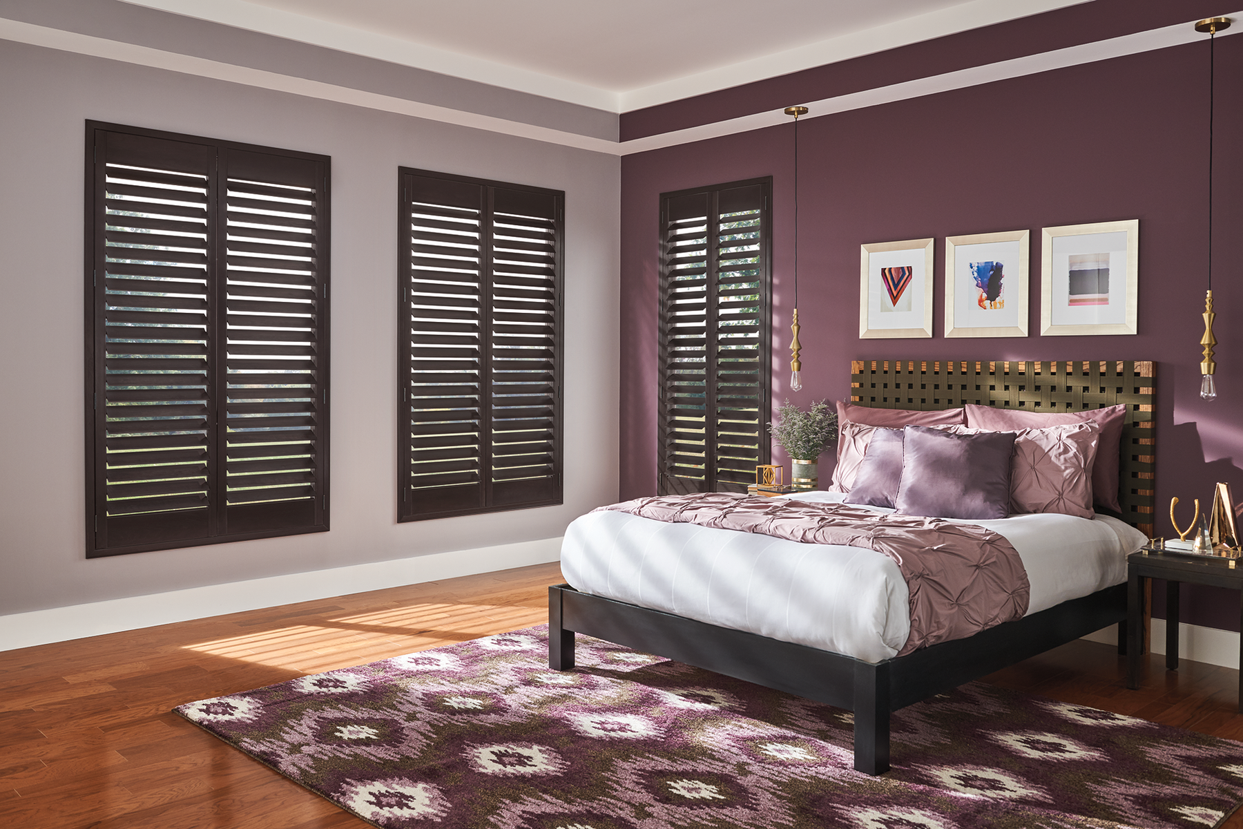 Dark wood shutters on three tall bedroom windows beside a bed with purple bedding