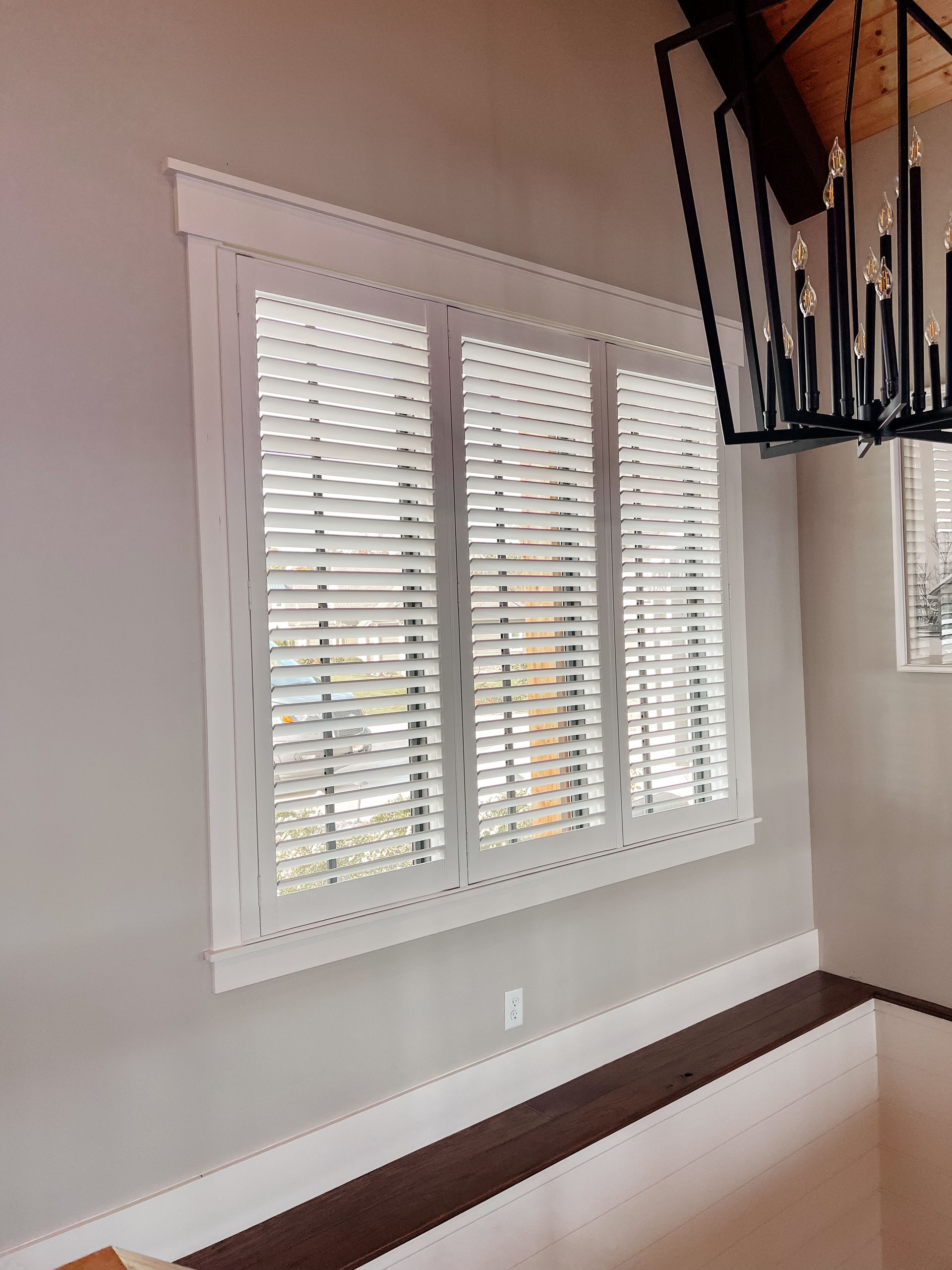 A dining room with a large window with shutters and a chandelier hanging from the ceiling.