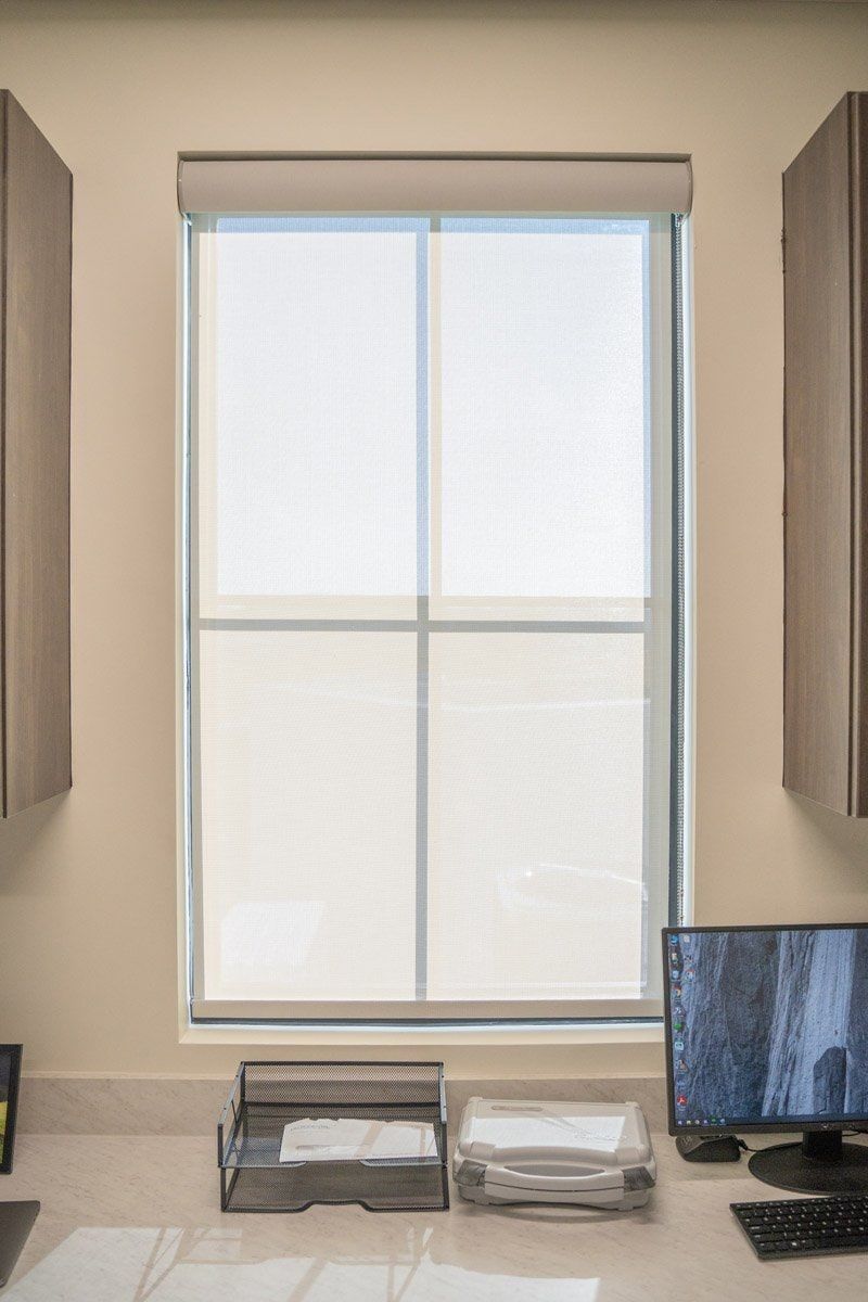A computer is sitting on a desk in front of a window with solar shades.
