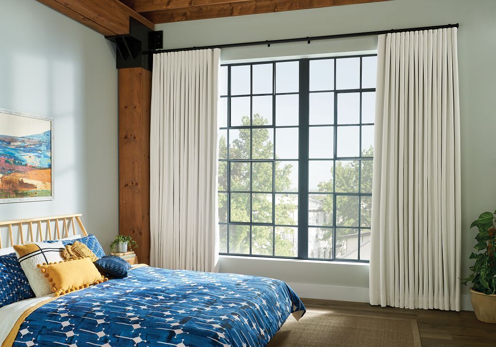 White pleated drapery framing a tall black-grid bedroom window above blue bedding