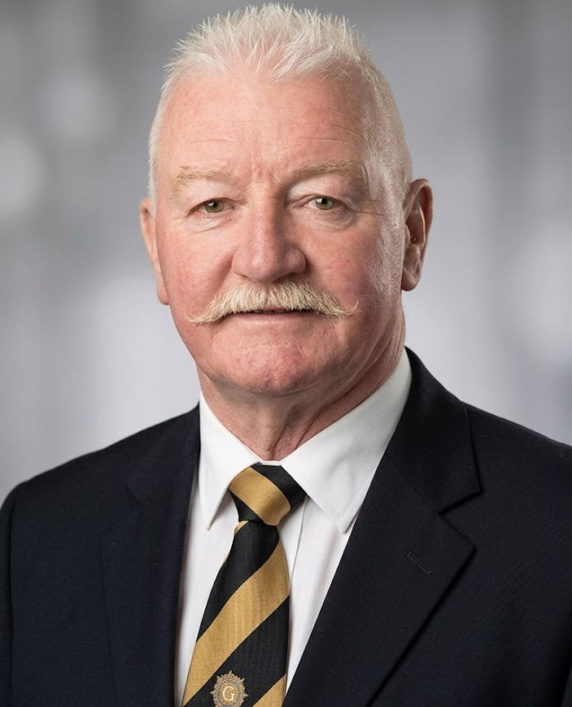 Portrait of a man in a dark suit with a black-and-gold striped tie, standing against a gray background