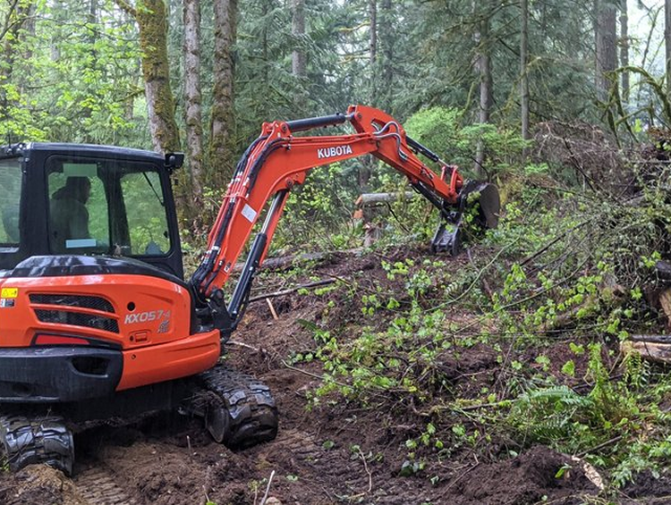 A small orange excavator is working in the woods.