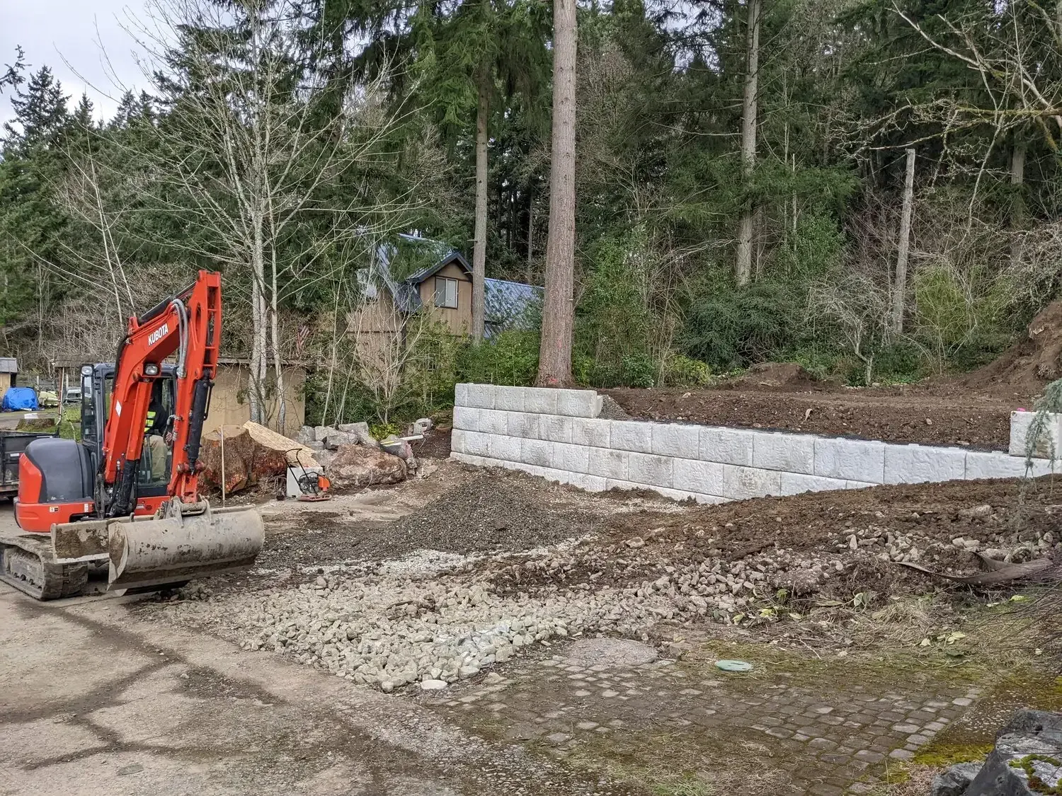A red excavator is parked in a dirt lot next to a stone wall.