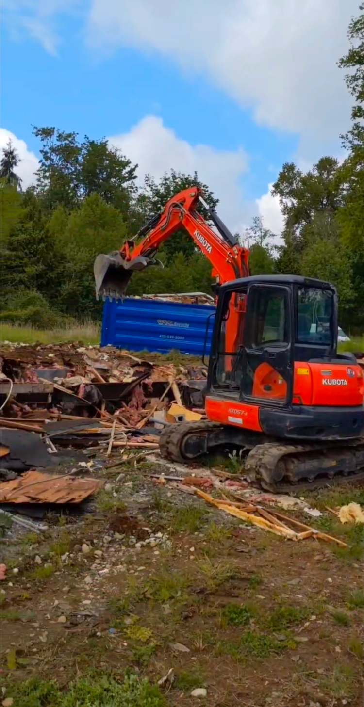 A red and black excavator is loading a blue dumpster with rubble.