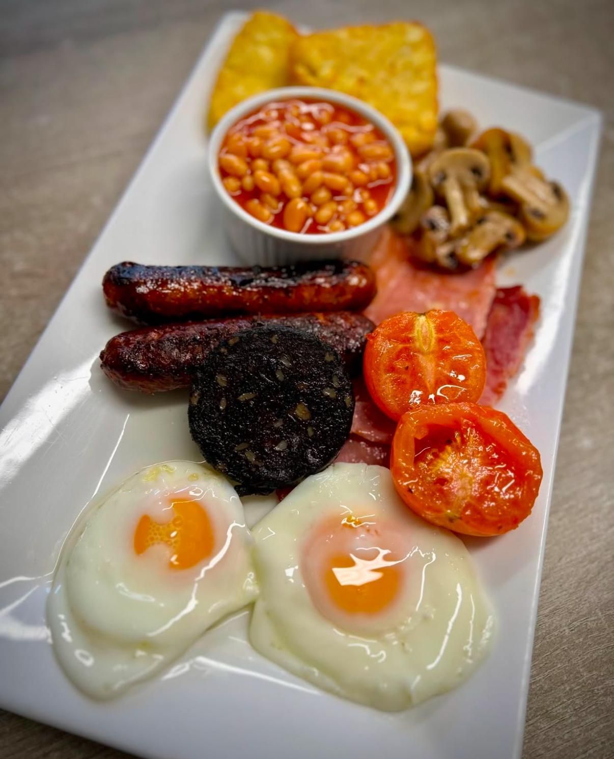 Full English breakfast: eggs, sausage, bacon, beans, mushrooms, tomatoes, hash browns, black pudding, on a white plate.