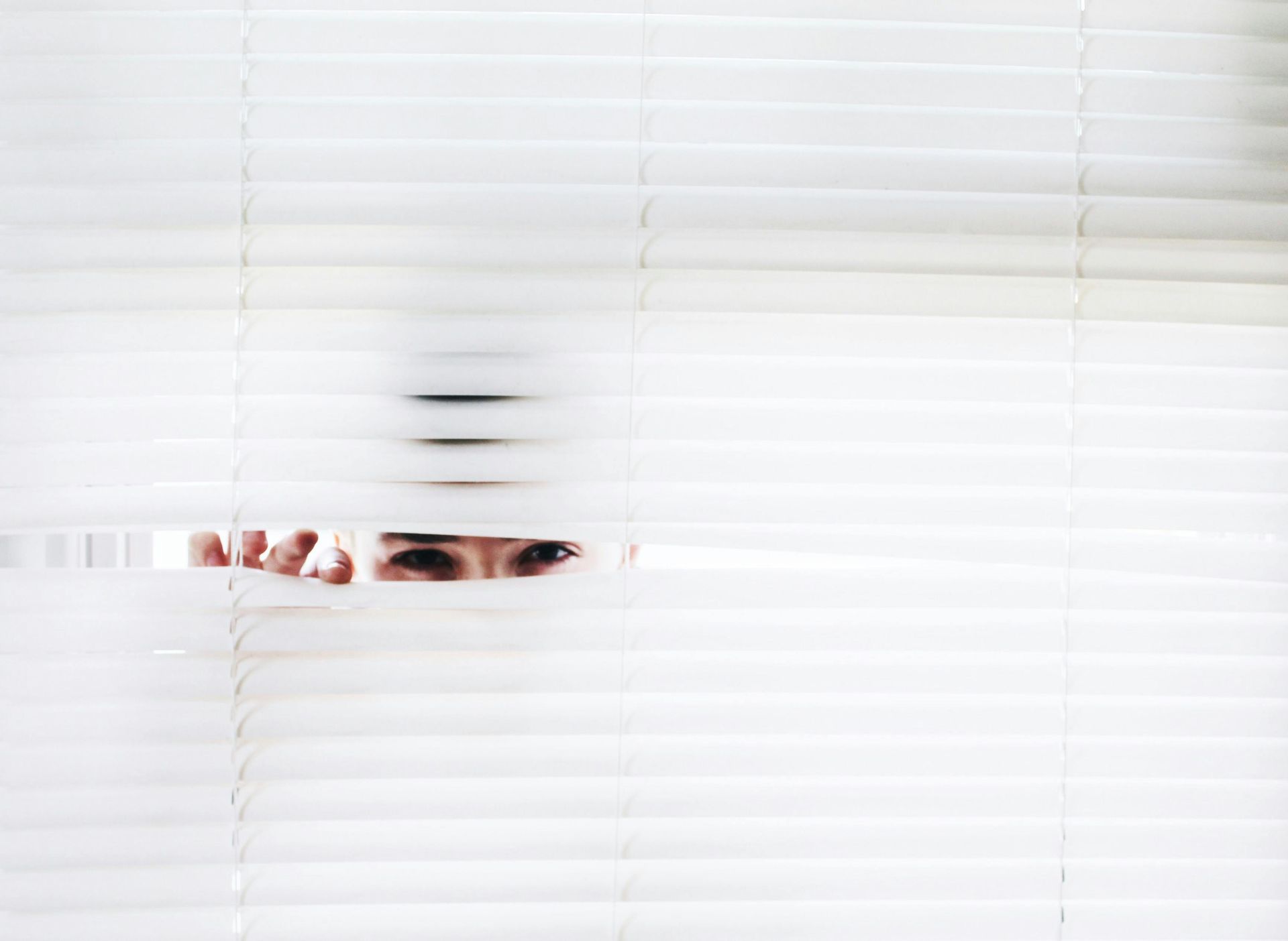 Person peeking through white blinds, eyes focused, hand visible.