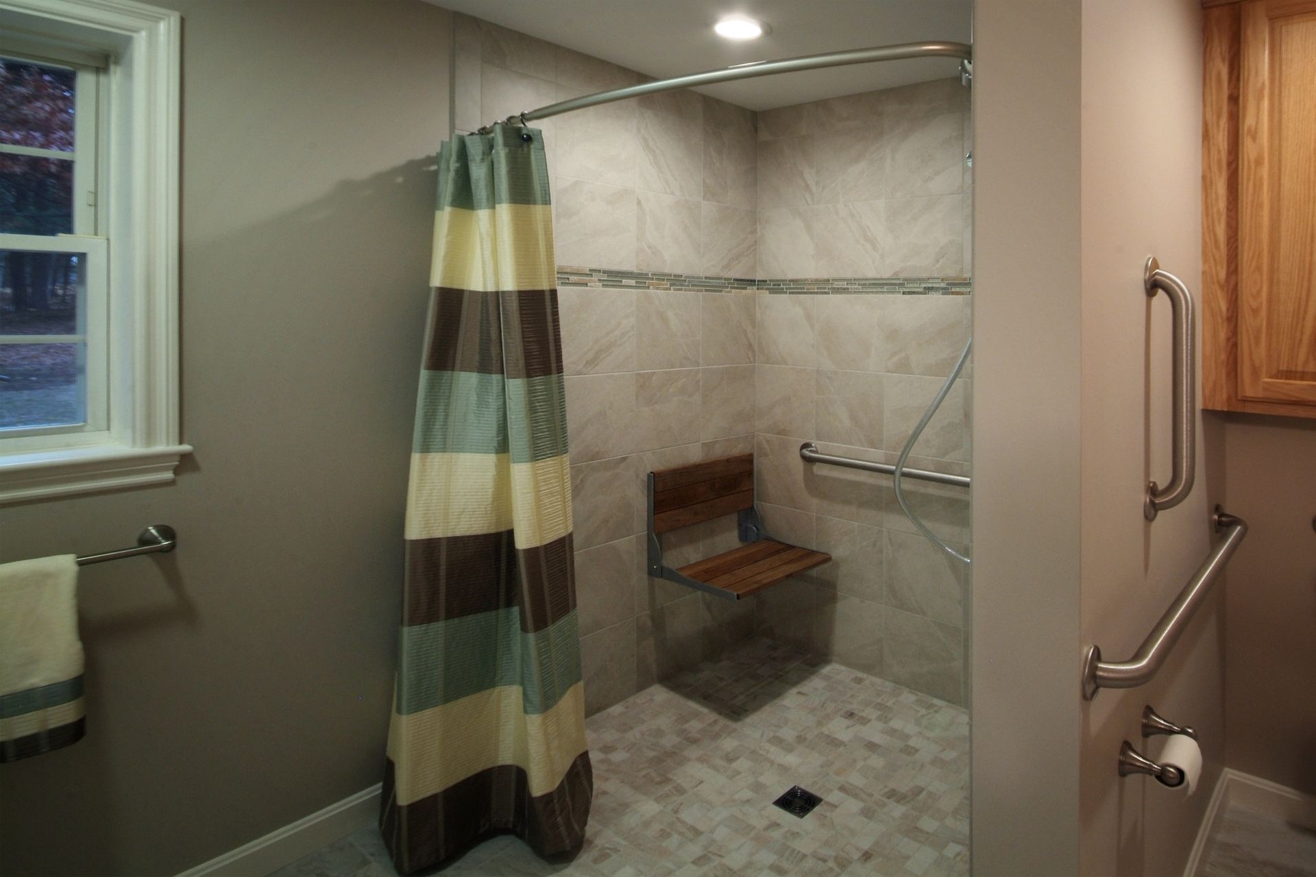 Bathroom with tiled walk-in shower, striped curtain, bench, window, and grab bars on the right wall