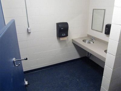 Empty public restroom stall with blue door, white tiled walls, and sink counter with mirror.
