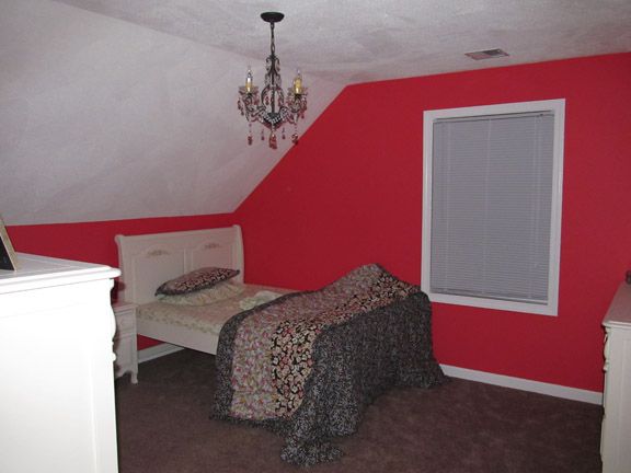 Small attic bedroom with red walls, white bed, chandelier, and a window with closed blinds