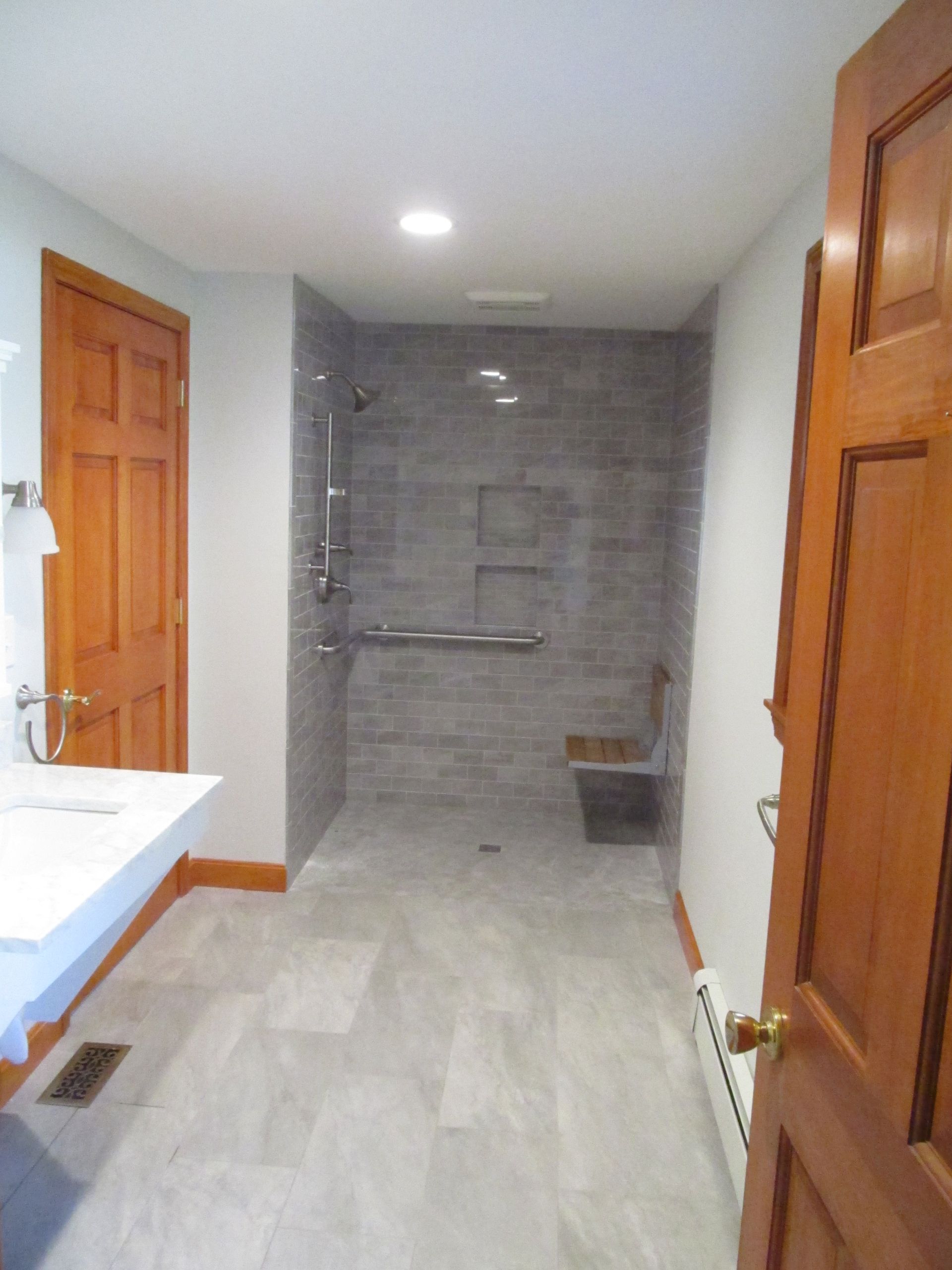 Bathroom with gray tiled shower, white vanity, and wooden doors in a narrow hallway