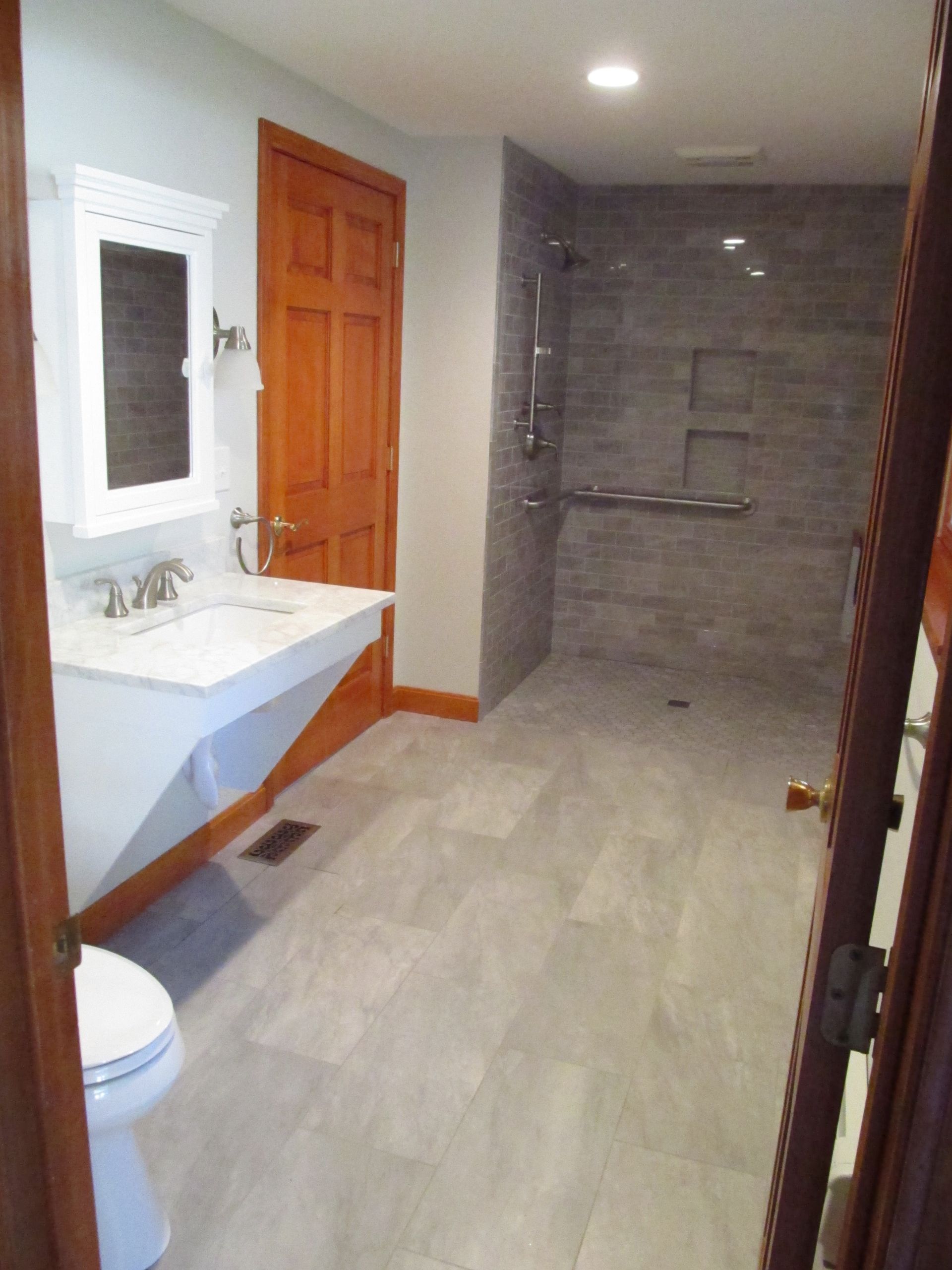 Modern bathroom with white vanity, wood door, tiled shower, and light gray flooring.