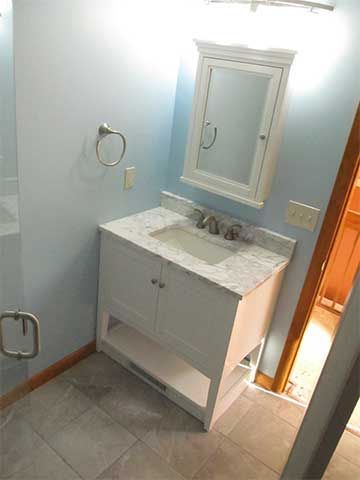 Bathroom vanity with white sink cabinet, mirror, towel ring, and tiled floor, seen from a doorway