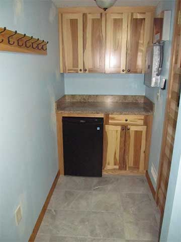 Narrow kitchenette with wood cabinets, black mini-fridge, countertop, and tiled floor in a small room