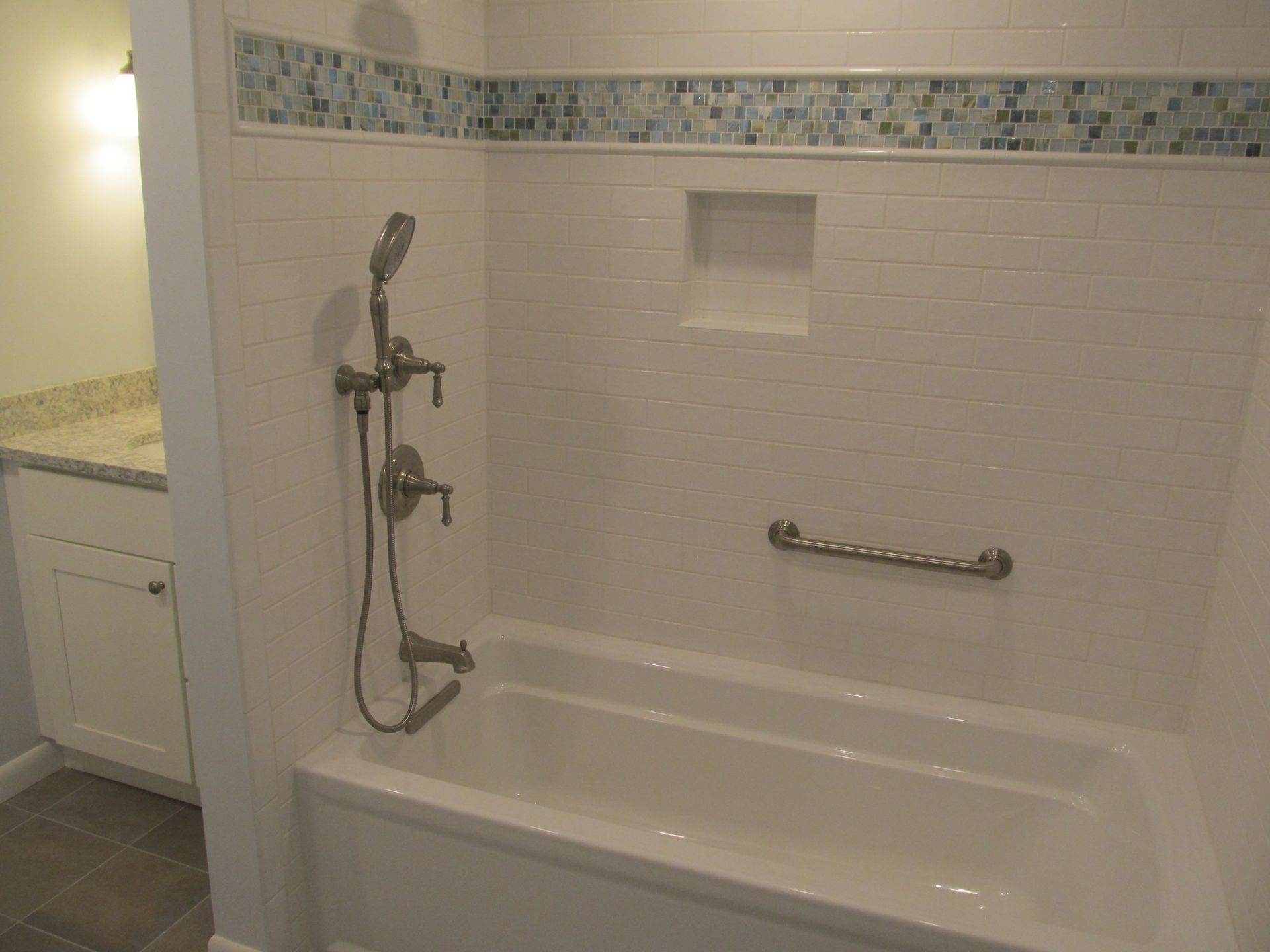 White tiled bathroom with a shower-tub combo, handheld shower, grab bar, and built-in niche.