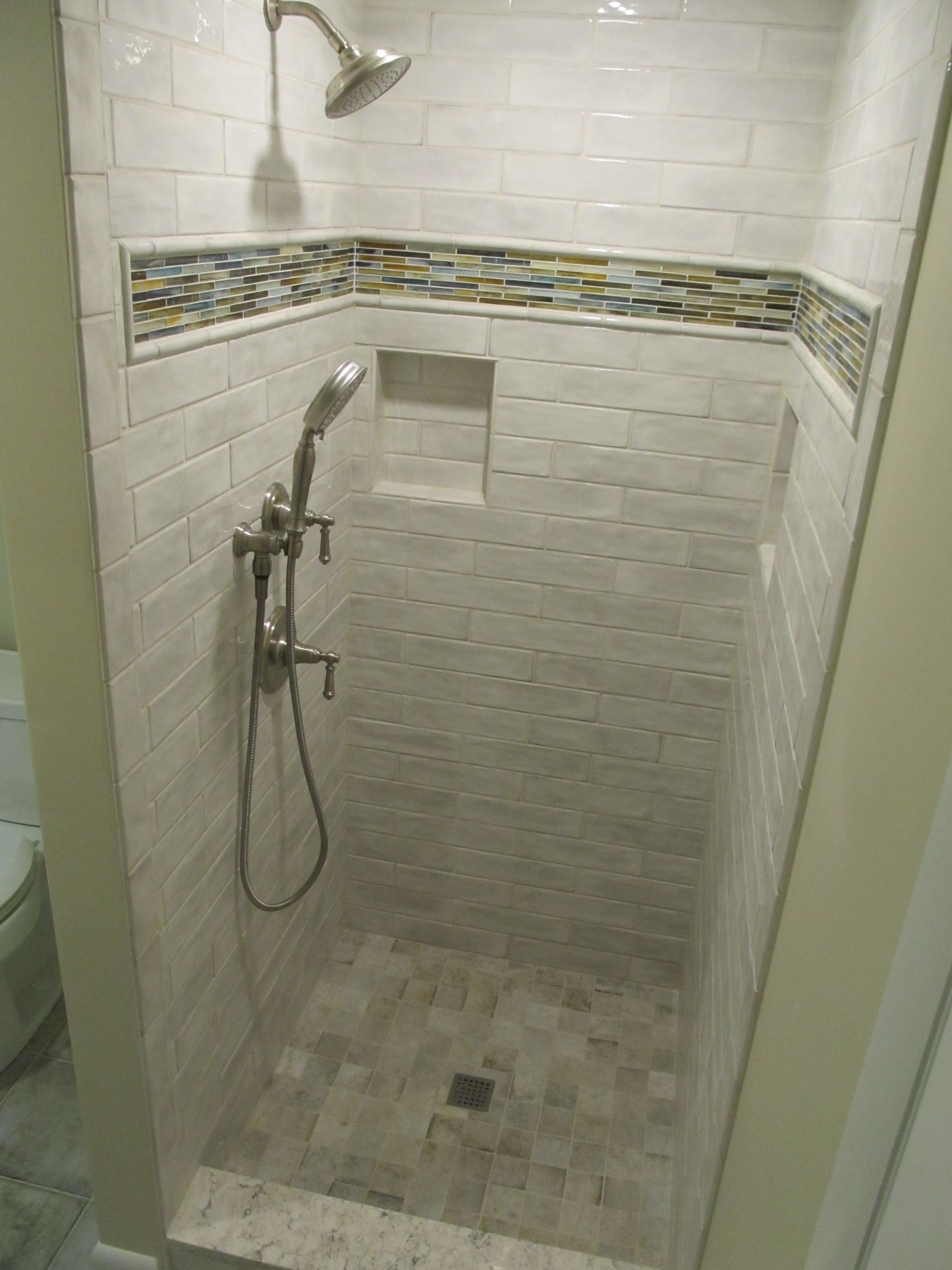 Small tiled shower stall with handheld showerhead, wall niche, and decorative border tiles.