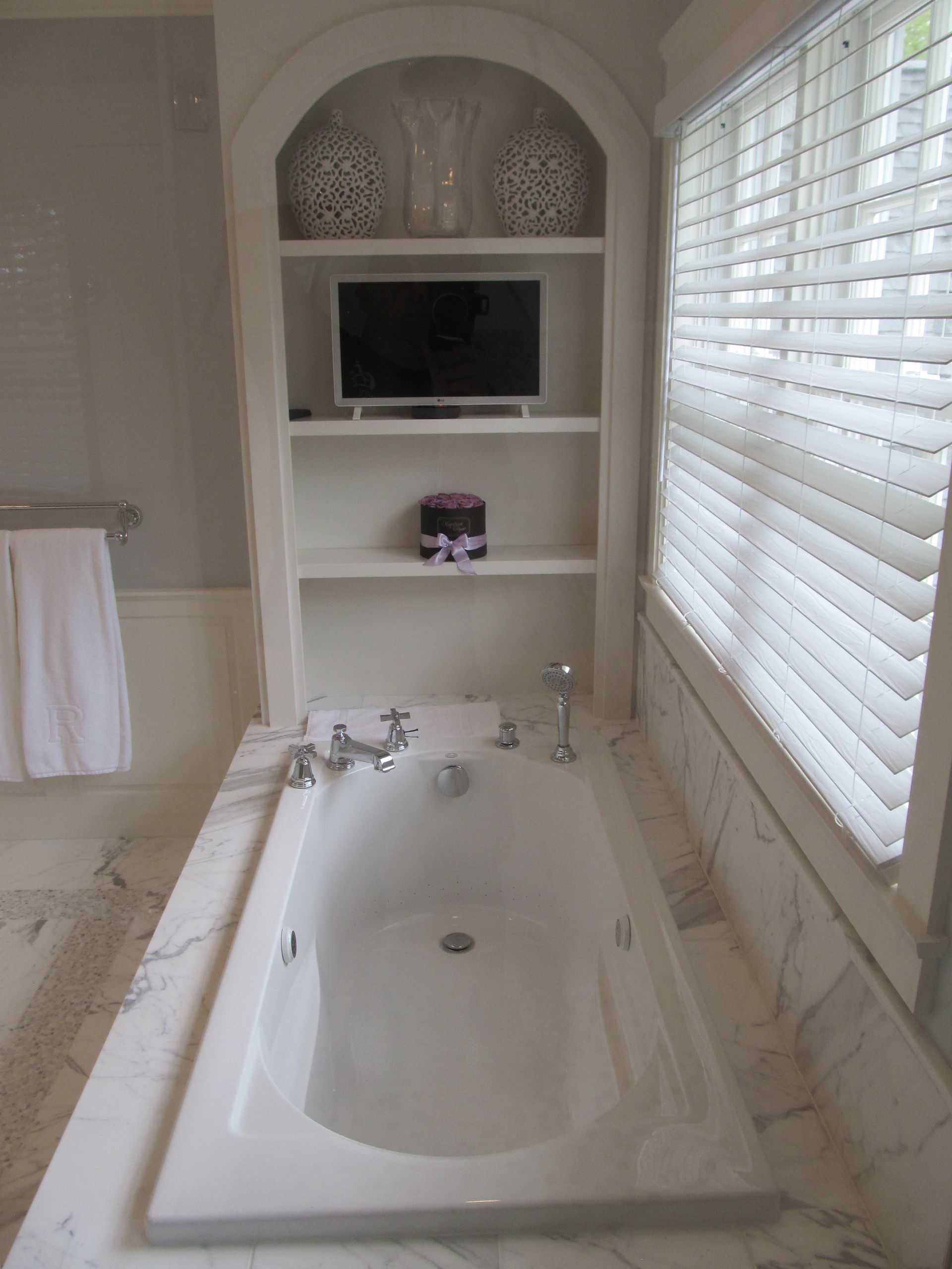 Bright bathroom with a soaking tub, built-in shelves, TV, and window blinds