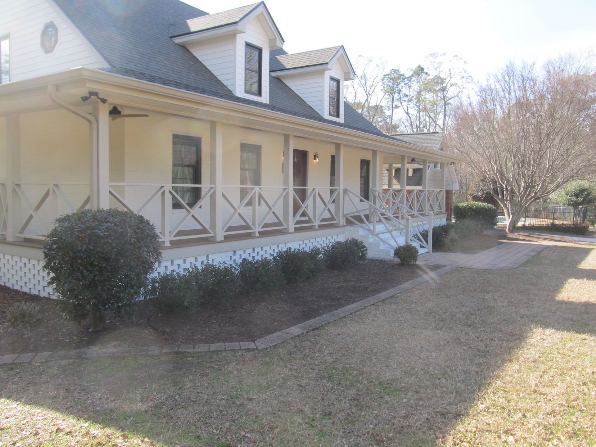 Large beige house with a covered porch, white railings, and dormer windows in a grassy yard