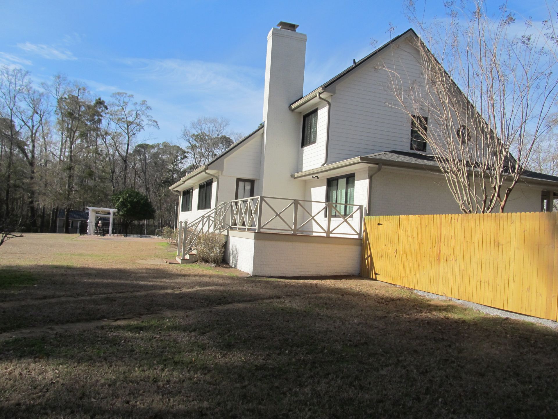 Suburban house with white siding, chimney, porch, and a wooden fence beside a bare yard and trees