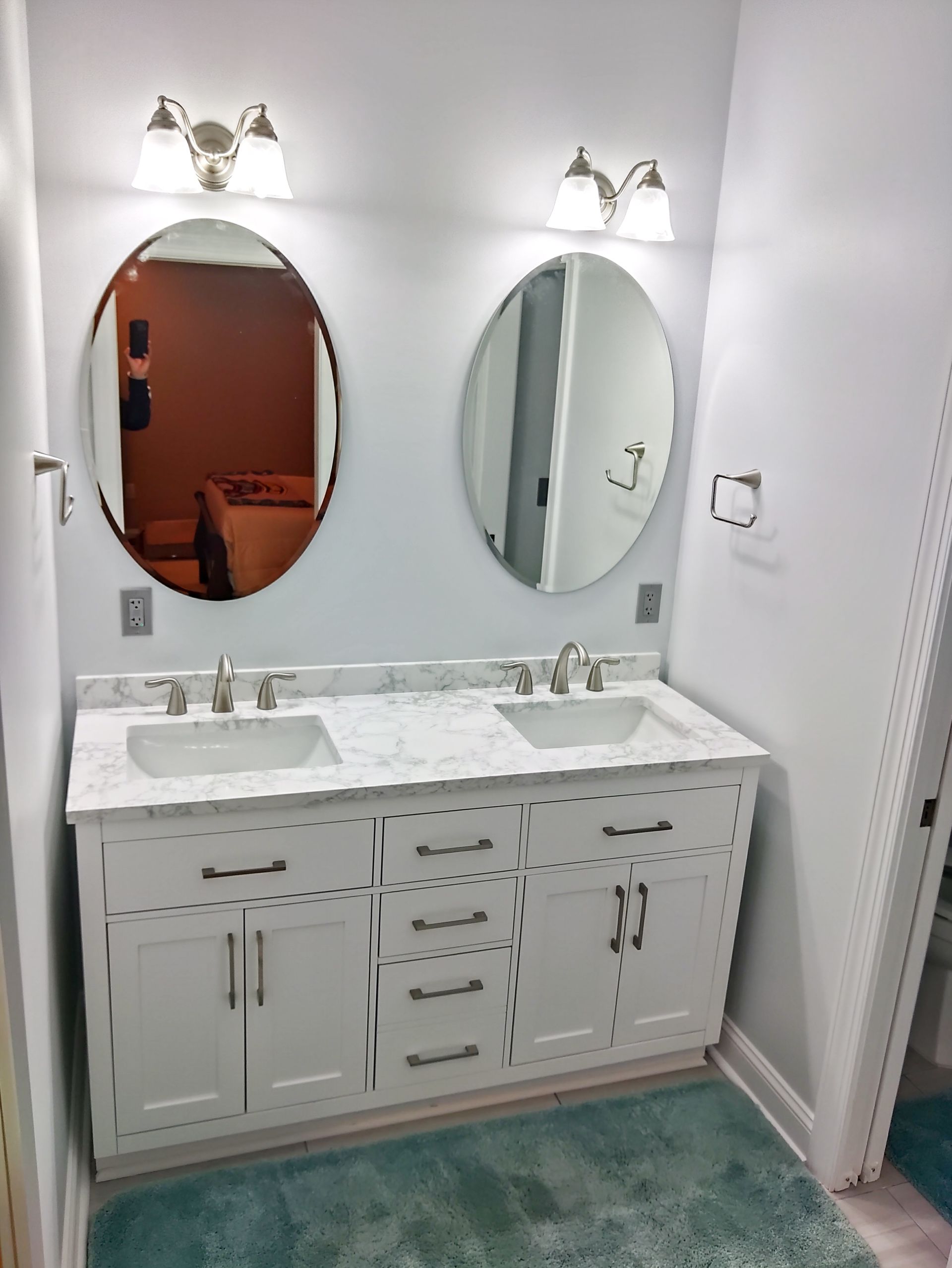 Double vanity bathroom with two oval mirrors, white cabinets, and teal rug.