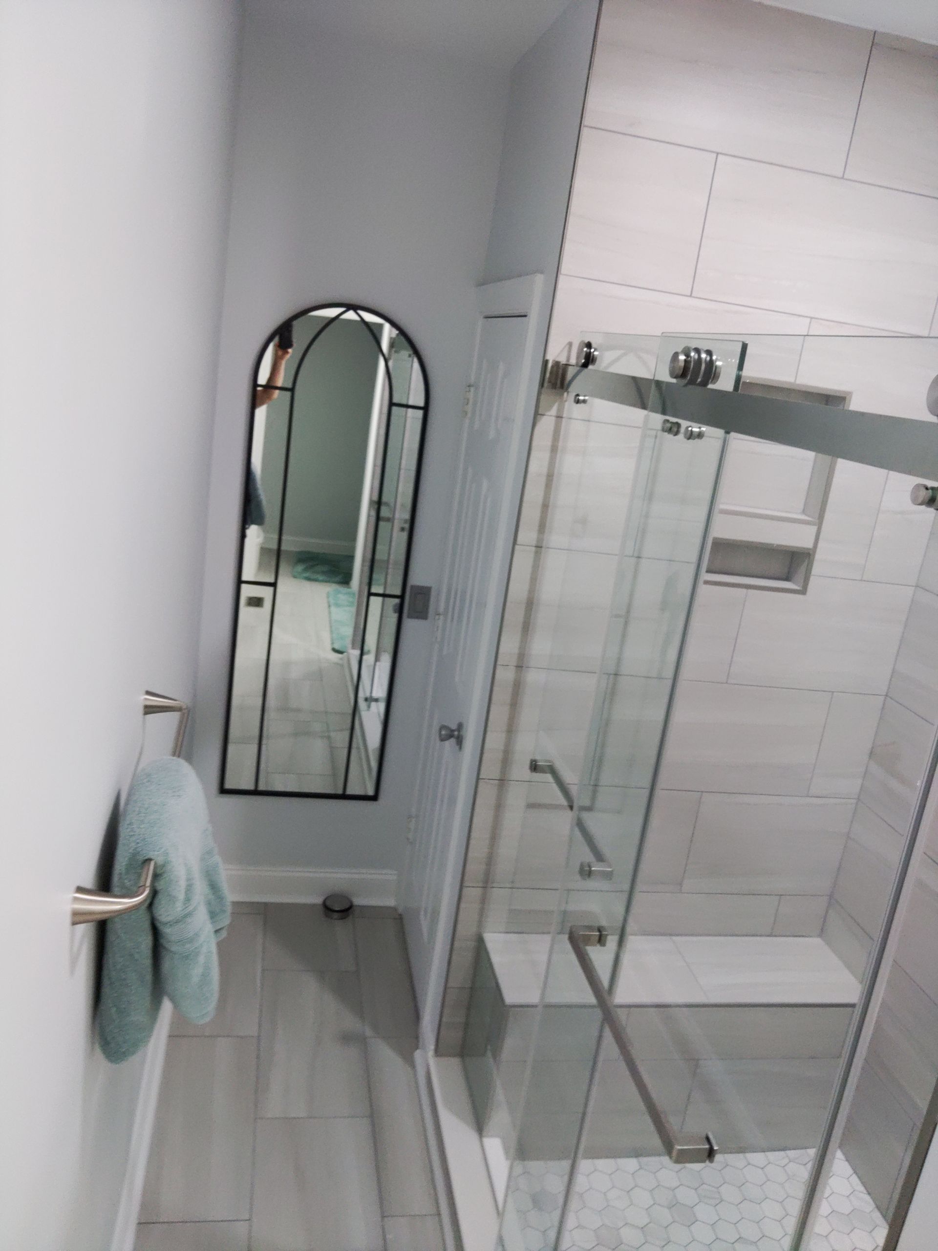 Narrow bathroom with glass shower, wall mirror, and blue towel hanging by the door