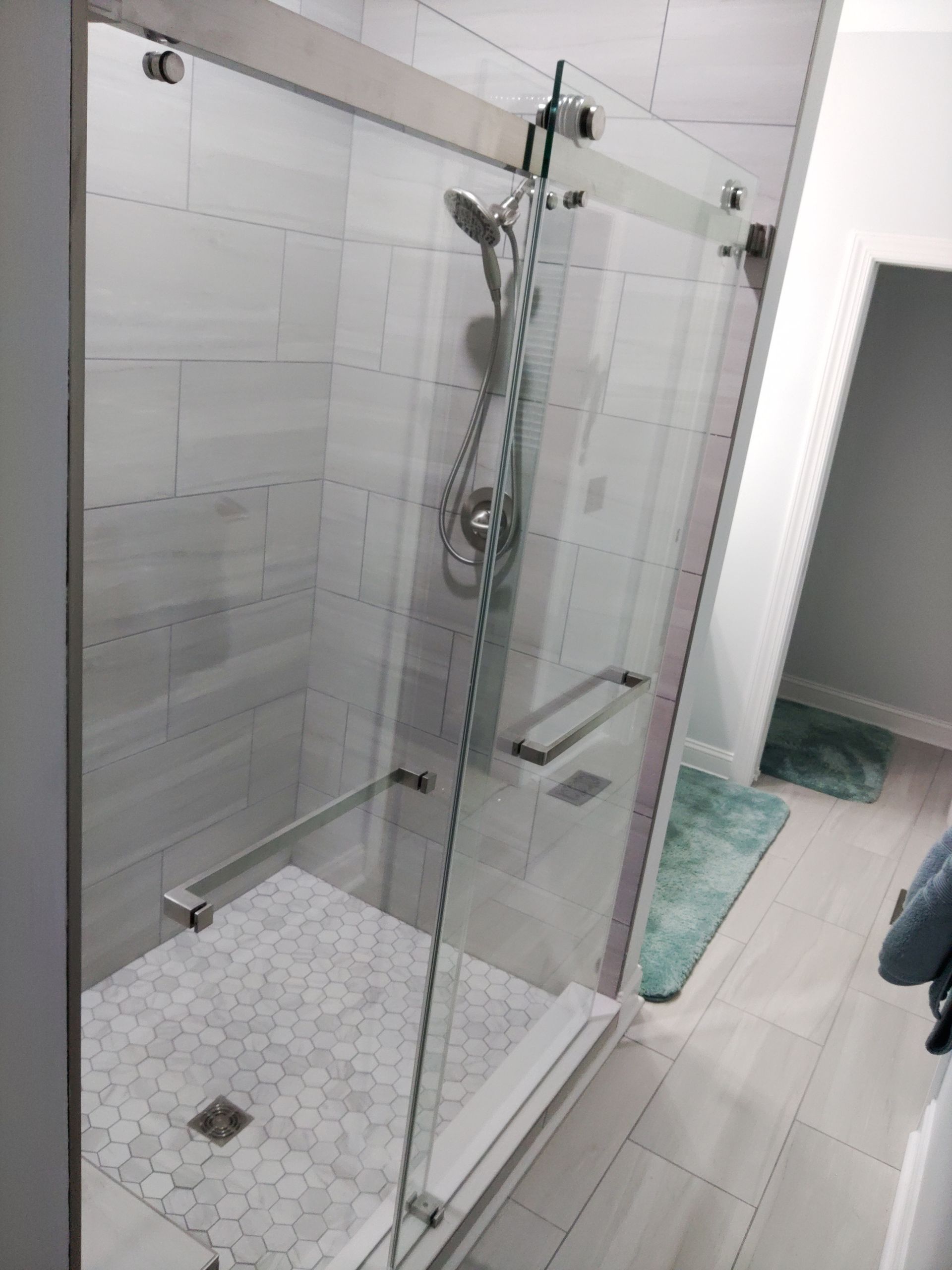 Modern bathroom with a glass walk-in shower, white tile walls, and a teal bath mat outside.