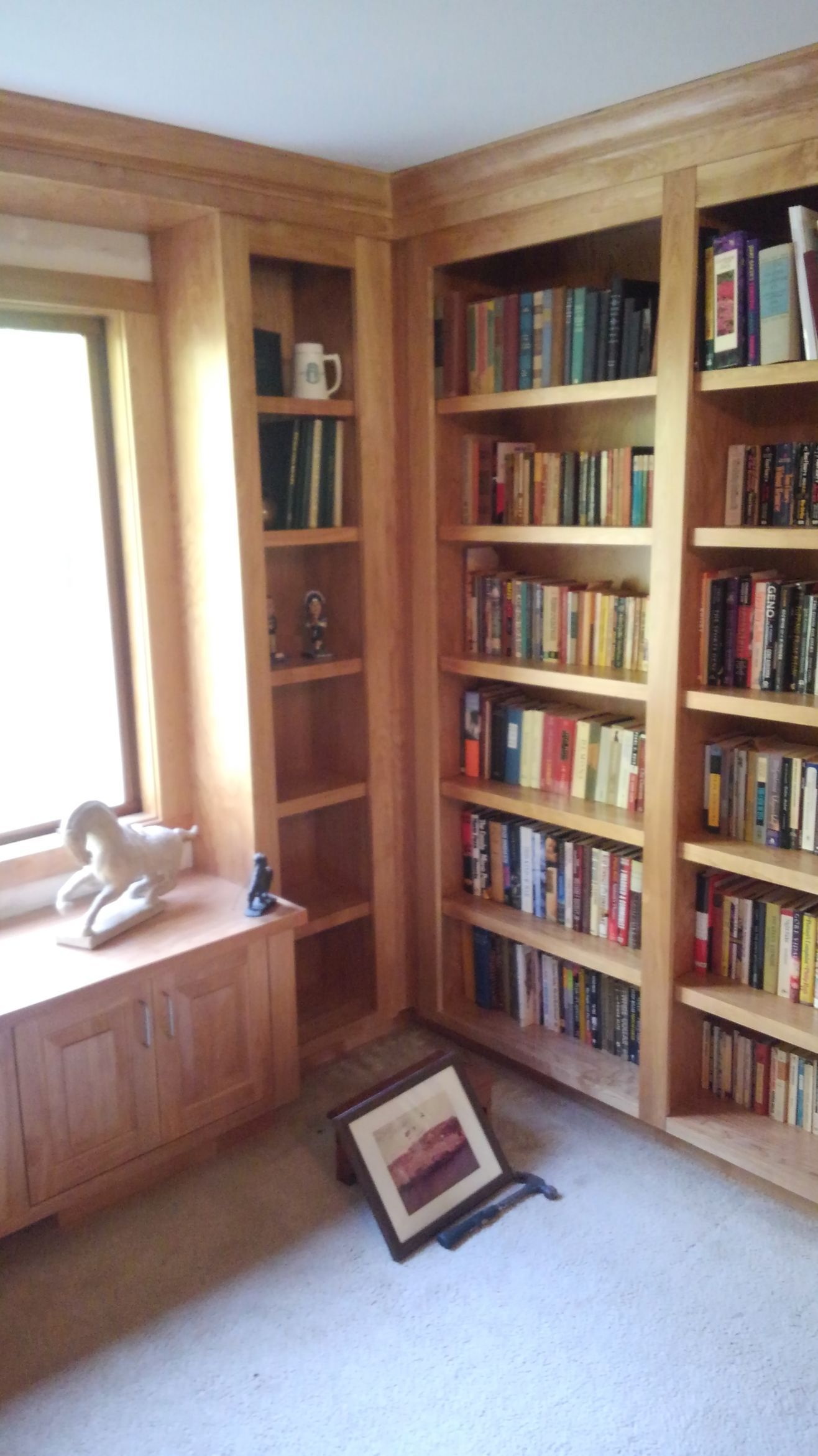 Built-in wooden bookshelves in a bright room, with books, a window seat, and a framed item on the carpet.