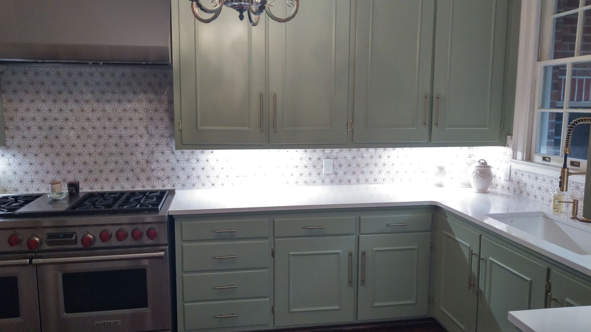 Bright kitchen with green cabinets, stainless stove, white countertop, and window above the sink.